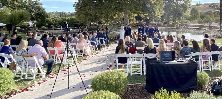 Wedding ceremony outside in front of a big tree near a pond. Guest sitting in white chairs on the lawn with live streaming video equipment and table set up in the back. Officiant about to invite the couple to kiss.